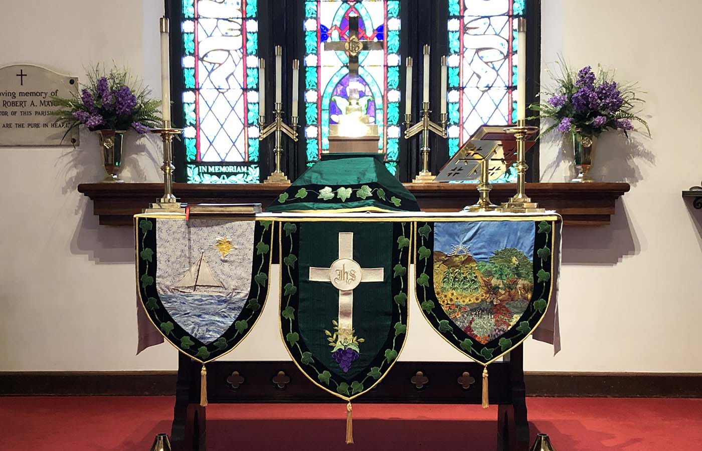 An intricately decorated altar with stained glass windows, green cloth banners, candles, and floral arrangements inside a church. Plaque dedicates altar in memory.