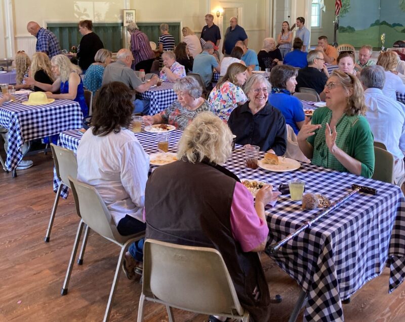 Hallcommunity A group of people socializing and eating at tables with checkered cloths inside a hall, featuring wood ceilings and a stage with artwork.