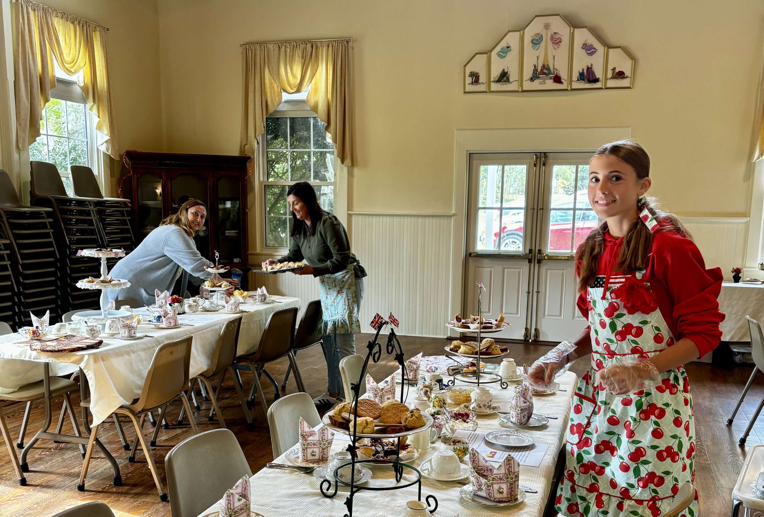 IMG_9604 Three people prepare an elegant tea party with treats and table settings in a bright room with large windows and cheerful decor.