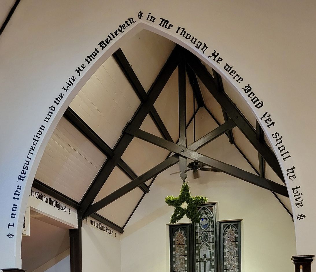 arch-sanctuary Arched ceiling with religious text, stained glass windows below, and a green wreath. Black metal beams support the structure, creating an architectural focus.
