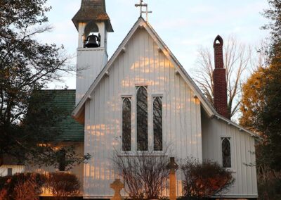 A small white church with a prominent steeple and cross, surrounded by gravestones, set amidst trees and bathed in warm light.