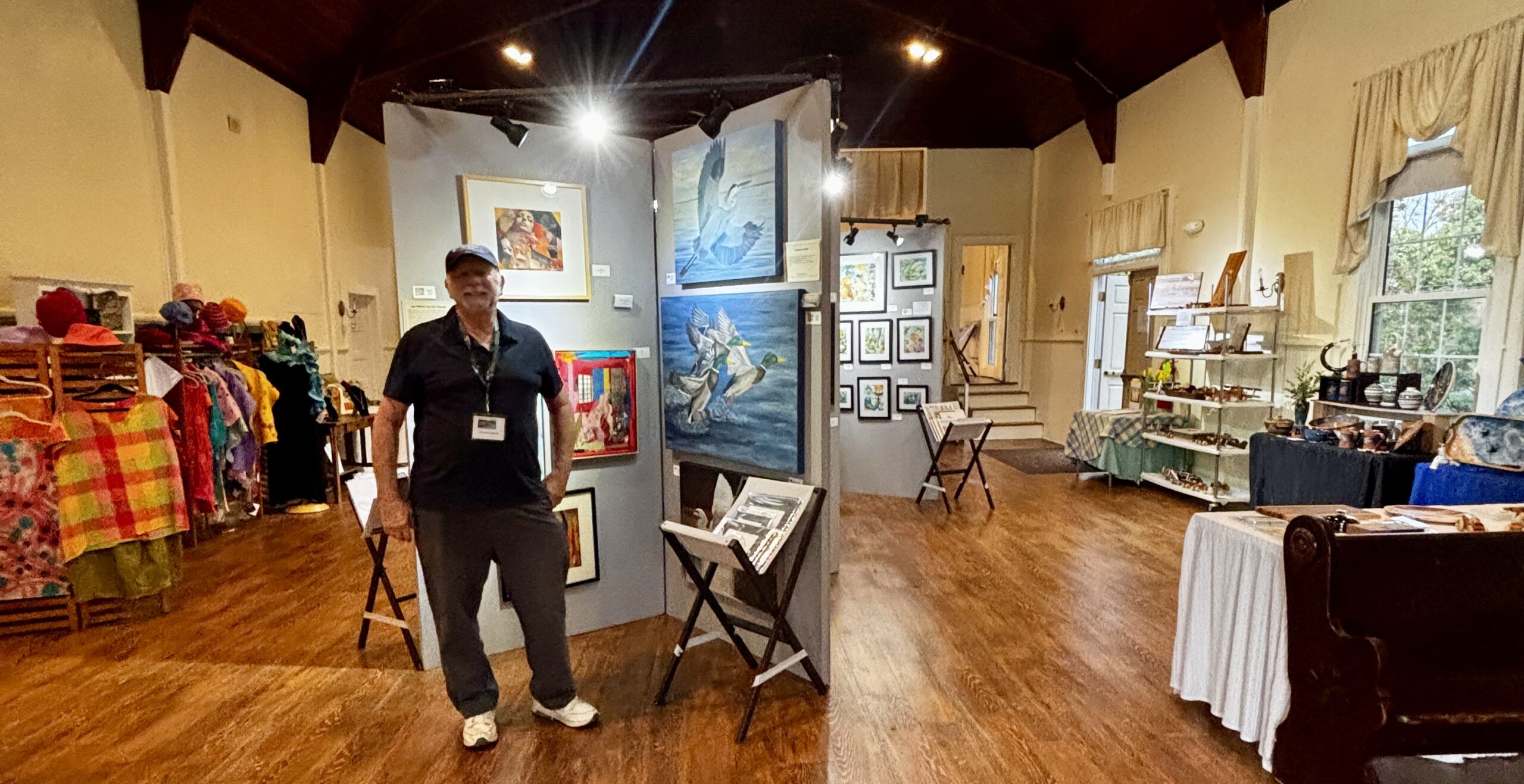 PH Art Show Layout jpg Art exhibition hall with paintings, crafts, and textiles. A person stands next to displayed artwork, wearing a badge. Bright, well-lit space.