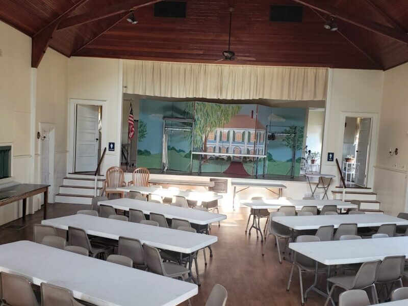 PH seminar central aisle angled tables setup 50 seated A hall with a wooden ceiling, white tables, and chairs faces a stage with an illustrated backdrop of a house and trees.