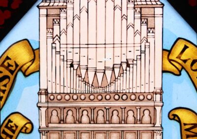 Stained glass artwork depicting a detailed pipe organ, surrounded by decorative banners with partial text visible, set against a light blue background.